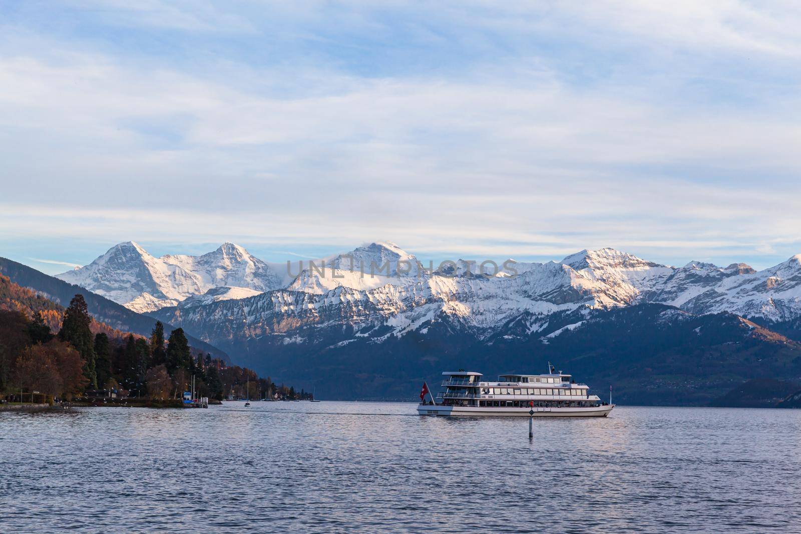 Stunning panorama view of famous Swiss Alps peaks on Bernese Oberland Eiger North Face, Monch, Jungfrau at dusk from Lake Thun (Thunersee) on sunny autumn day with cruise ship, Bern, Switzerland