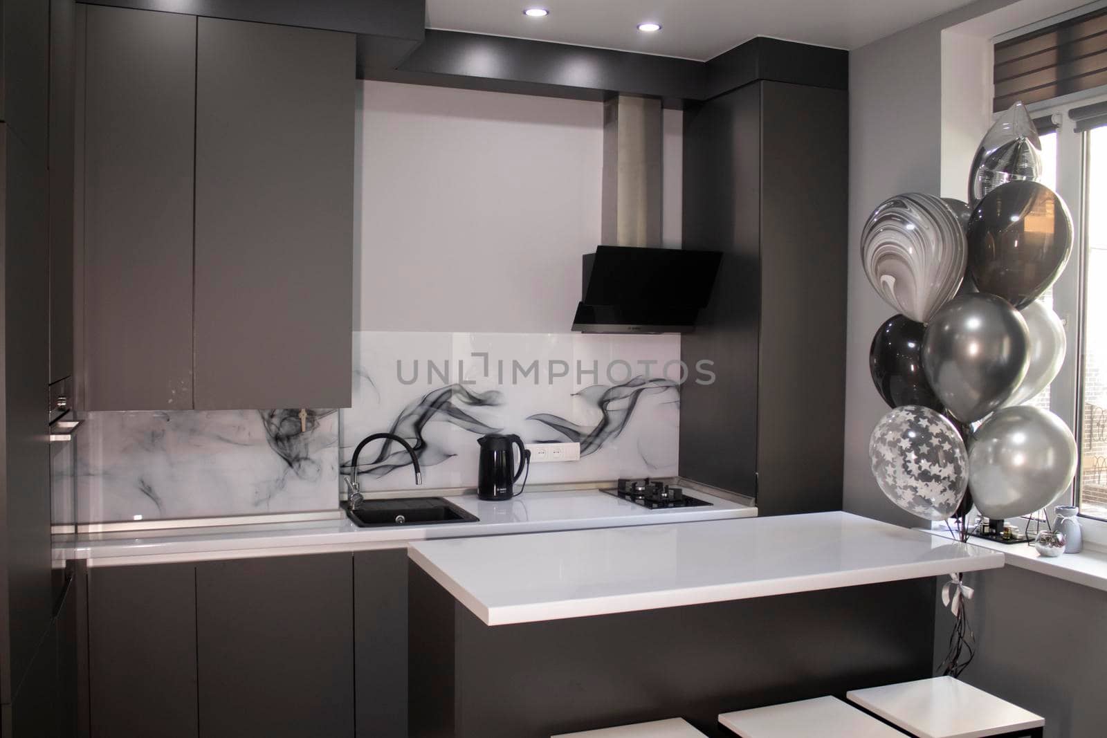 Corner of stylish Minimalistic grey kitchen, dark gray cupboards, white countertops and bar with stools. black sink, kettle and stove. and black and white balloons. High quality photo