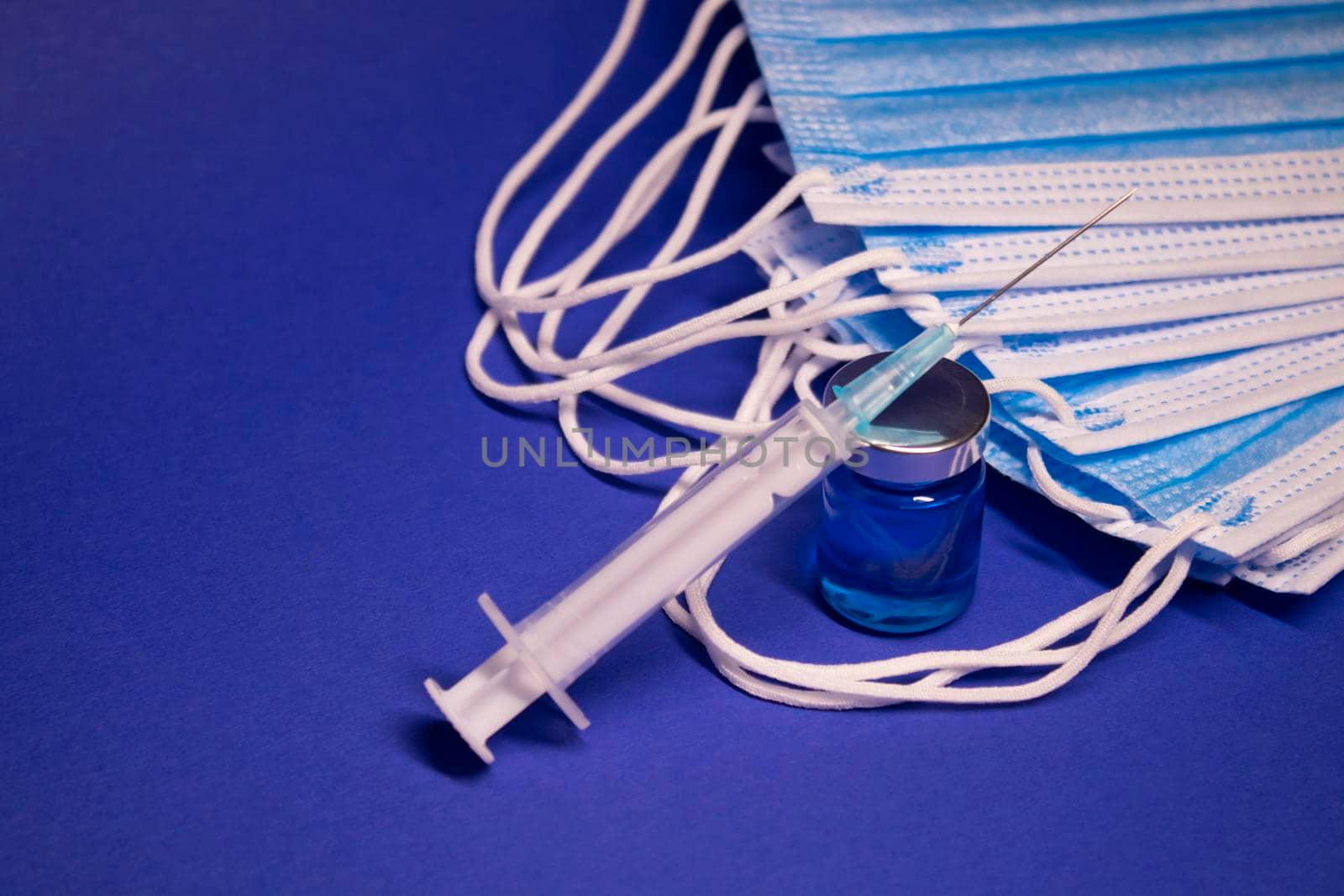 Vaccine bottle phial with no label and medical syringe with injection needle near pile of blue medical masks . isolated on blue background. cure. Development of coronavirus vaccine COVID19. copy space