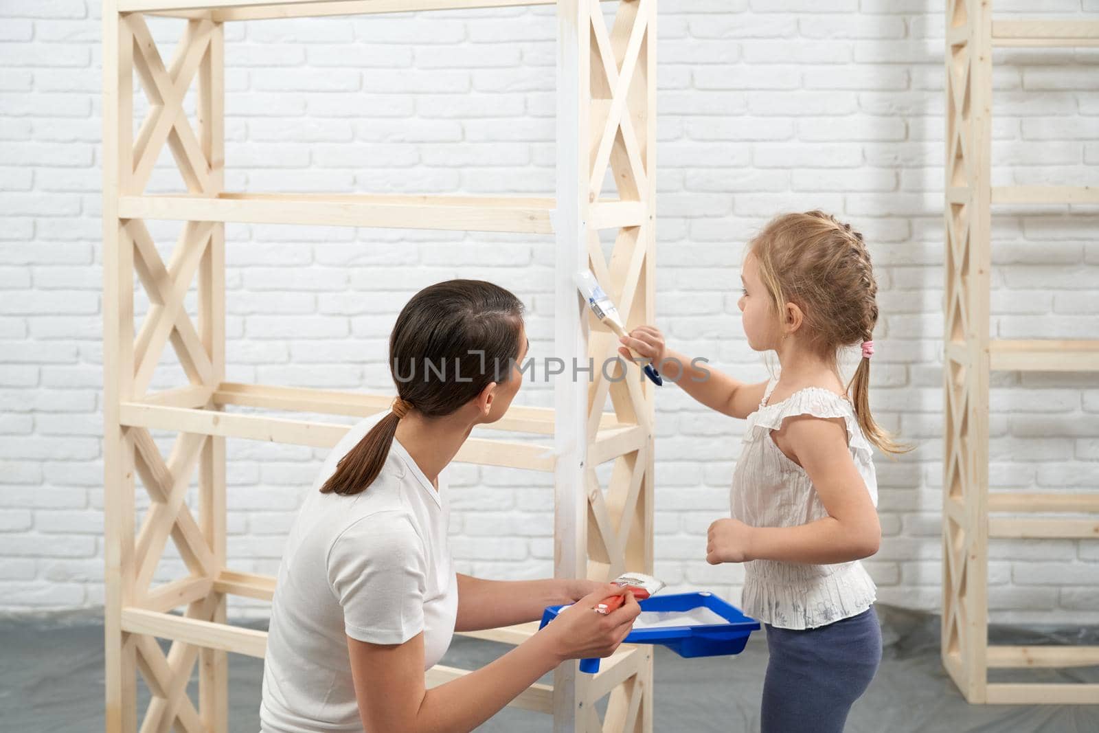 Young brunette woman holding white paint and child painting wooden rack at home. Concept of process painting mother and daughter.