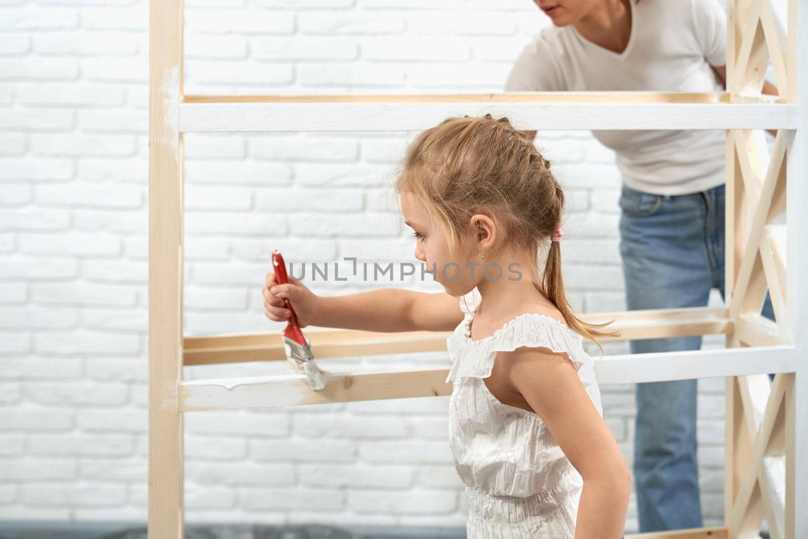 Mother and daughter together painting wooden rack in white color at home. Concept of family time and repair at home.