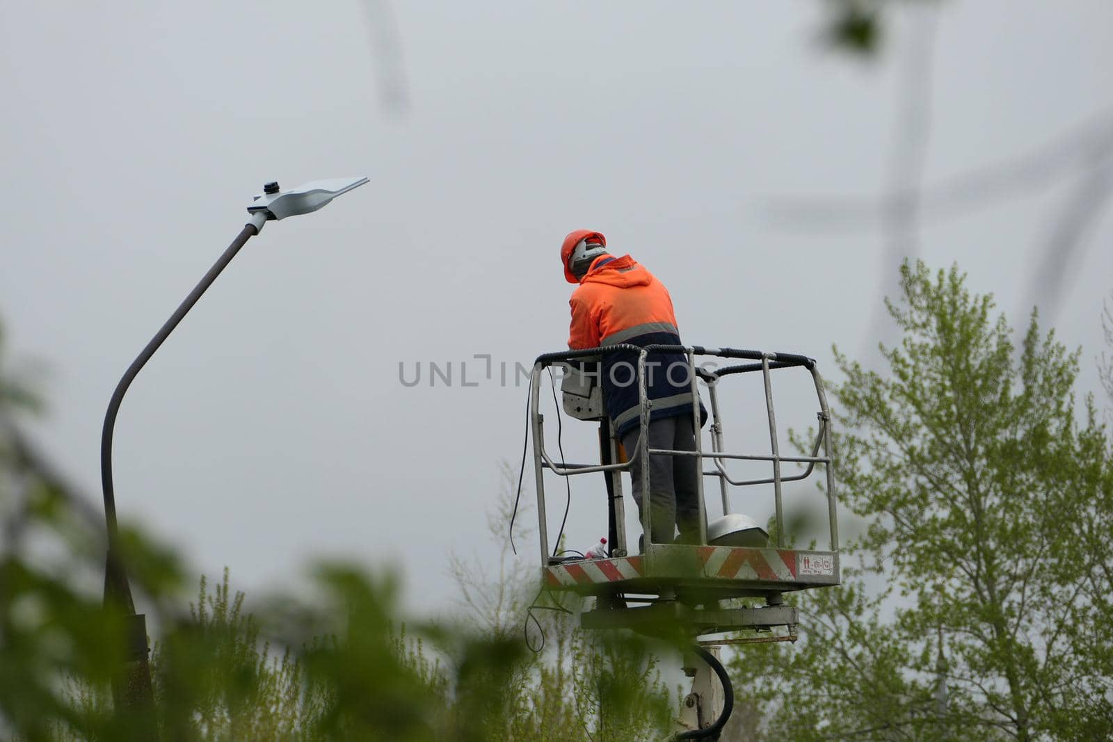 An electrician in special clothing and a helmet repairs a street lamp and screws a street light bulb on a special lifting platform. by Olga26