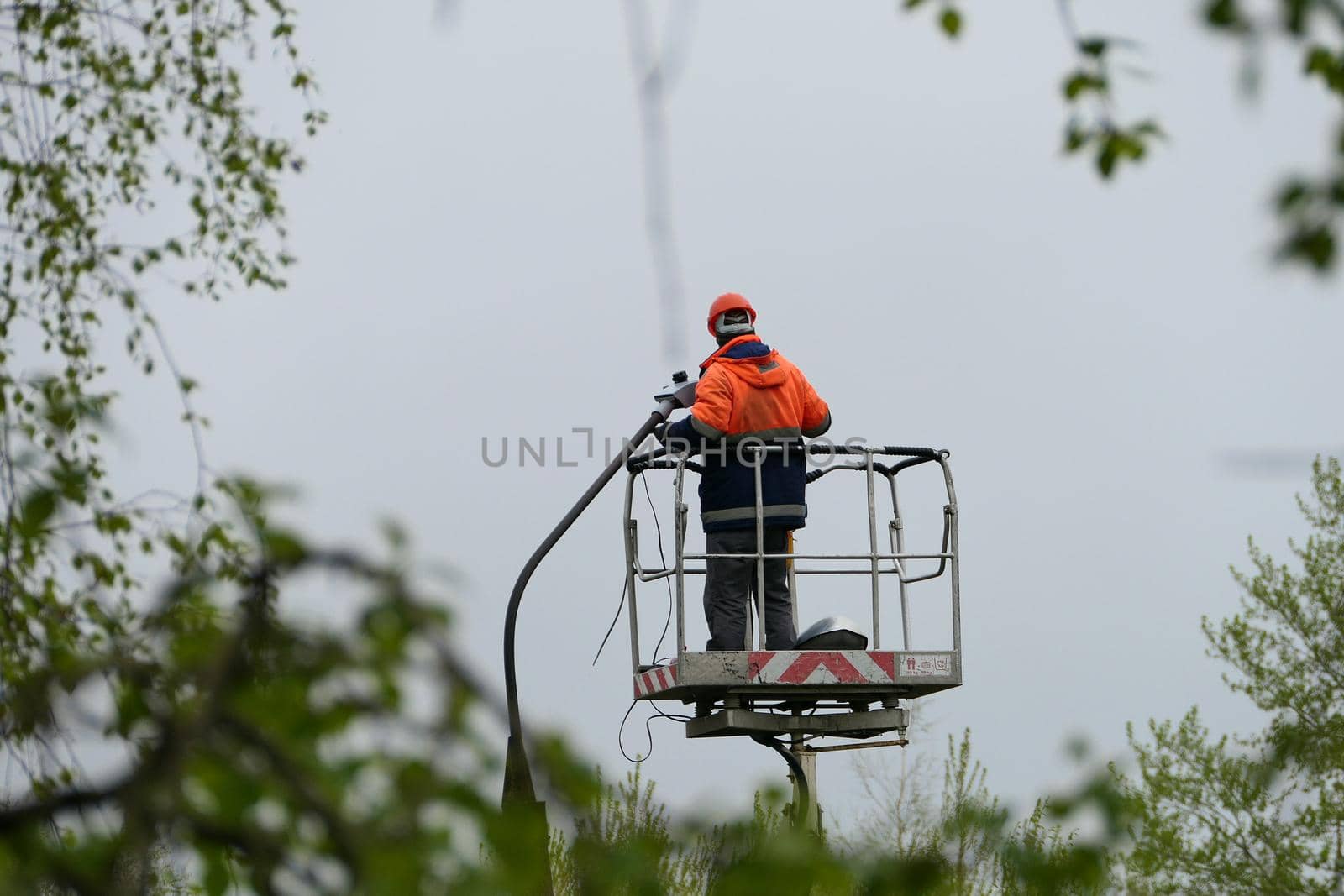 An electrician in special clothing and a helmet repairs a street lamp and screws a street light bulb on a special lifting platform. High quality photo