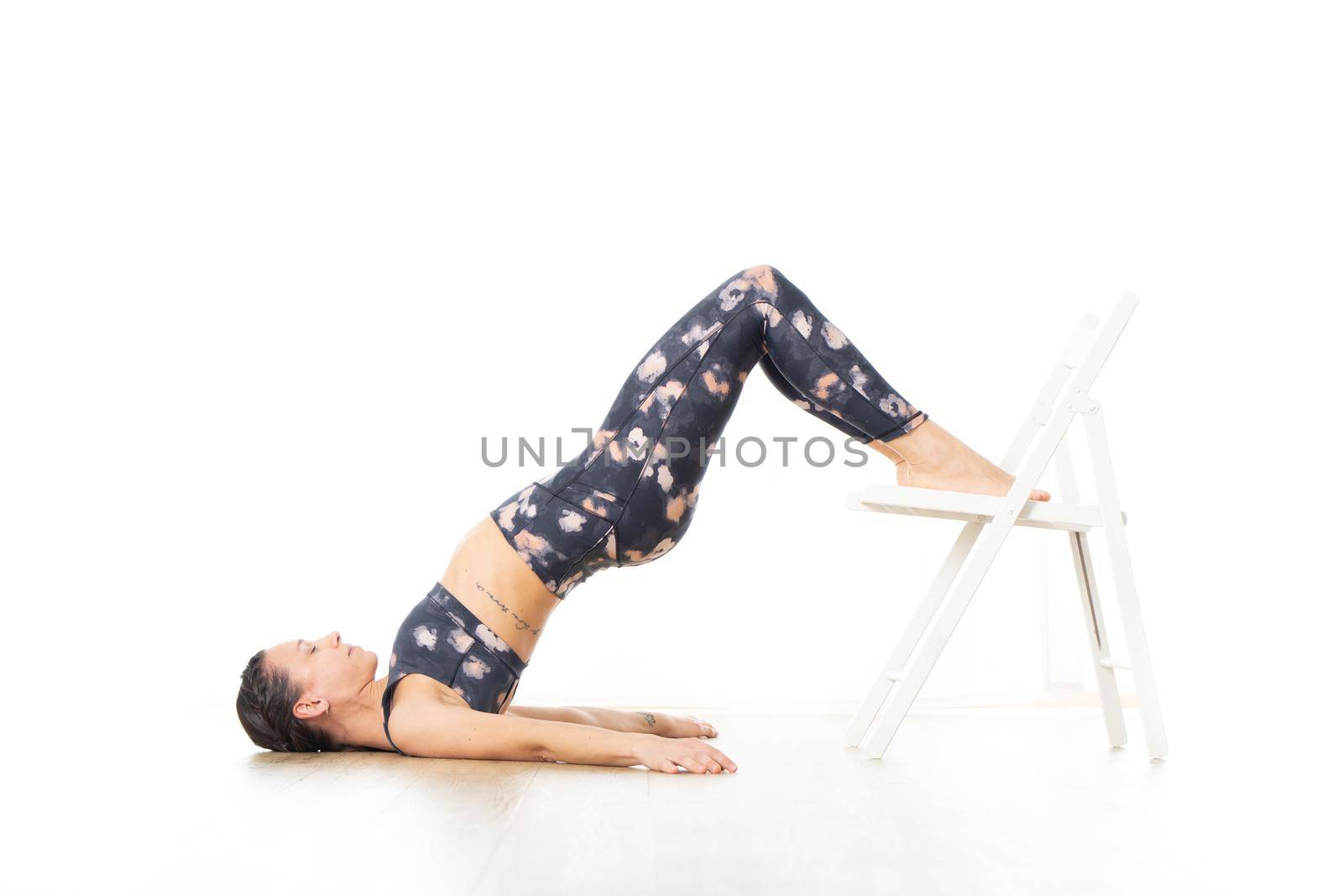 Young sporty attractive woman in bright white yoga studio, stretching and relaxing during restorative yoga using white chair as a gadget. Healthy active lifestyle by kasto