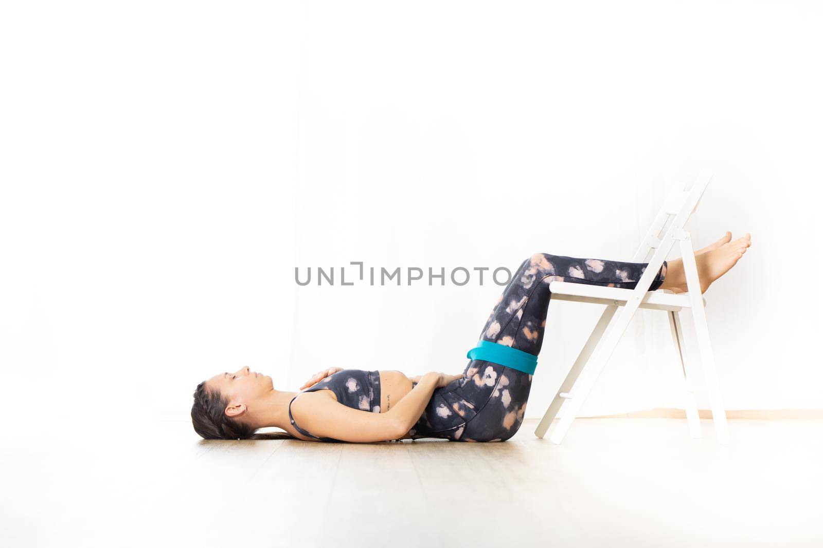 Young sporty attractive woman in bright white yoga studio, stretching and relaxing during restorative yoga using white chair as a gadget. Healthy active lifestyle by kasto