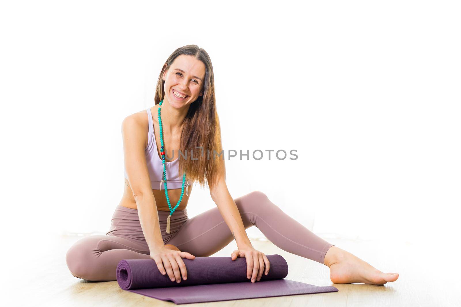 Young sporty female yoga instructor in bright white yoga studio, smiling cheerfully while rolling the yoga mat after yoga session. by kasto