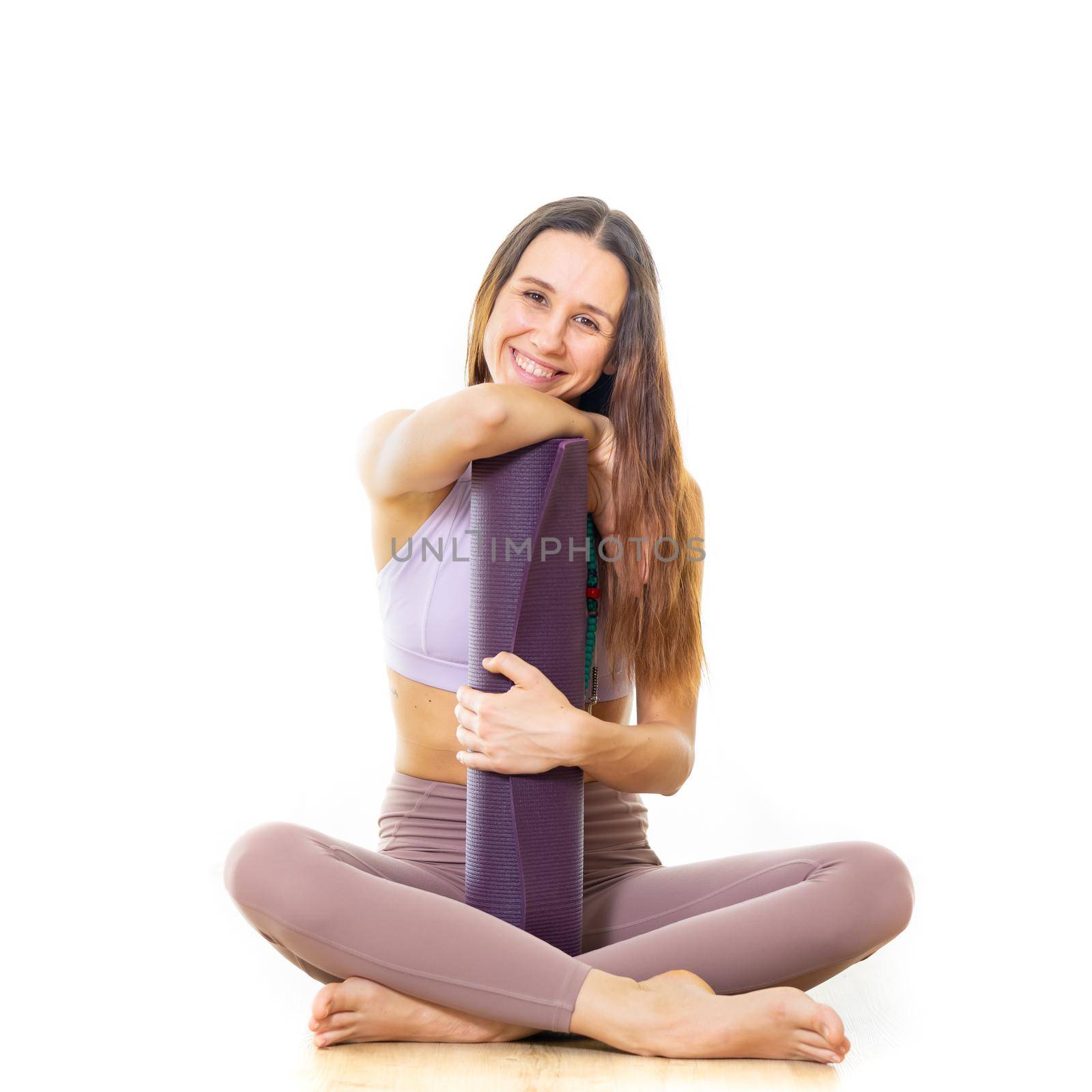 Young sporty female yoga instructor in bright white yoga studio, holding yoga mat, smiling, looking at camera.