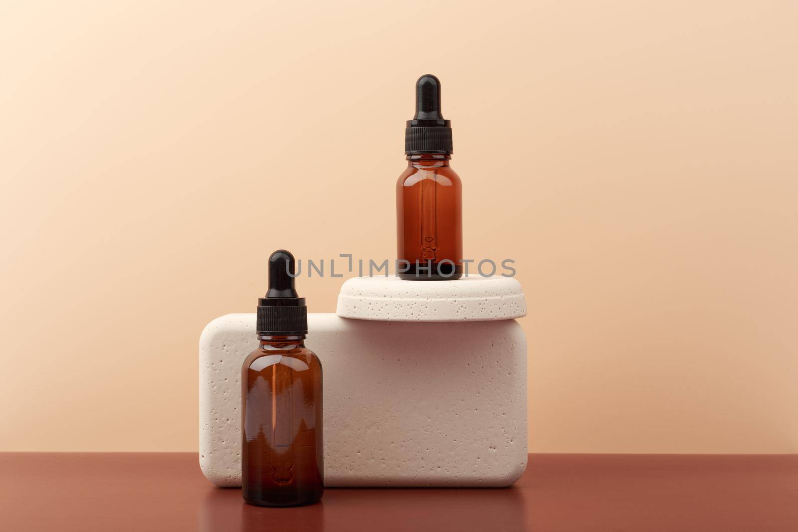 Two dark bottles with skin serum or oil for nails with beige geometric props on brown table against beige background with copy space. Concept of skin care or manicure
