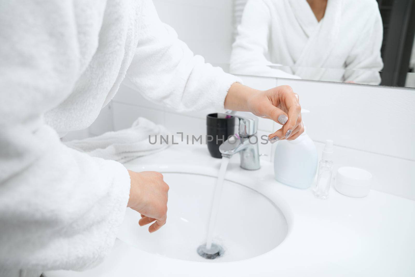 Side view of young woman in white robe standing near sink and washing hands with special antibacterial soap at home. Concept of body procedure for improvements skin.