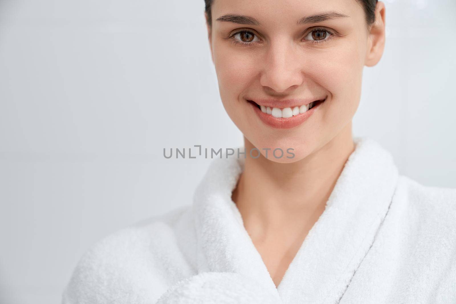 Close up portrait of smiling attractive woman in white robe standing in bathroom after shower. Concept of free time with useful procedures for body and improvements skin at home.