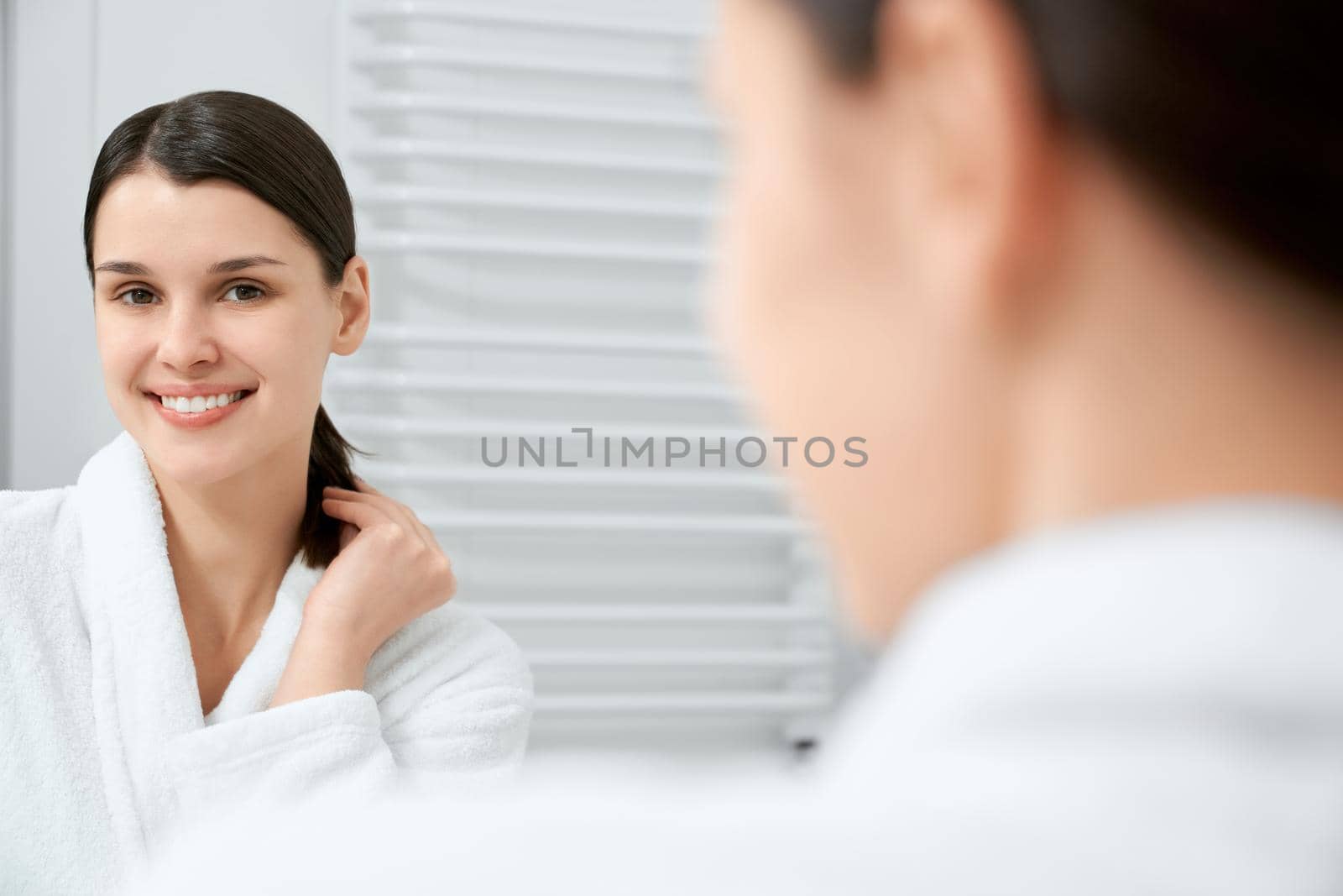 View from shoulder of smiling young beautiful woman standing in bathroom after shower and looking at mirror. Concept of relaxing time after beauty procedure at home. 