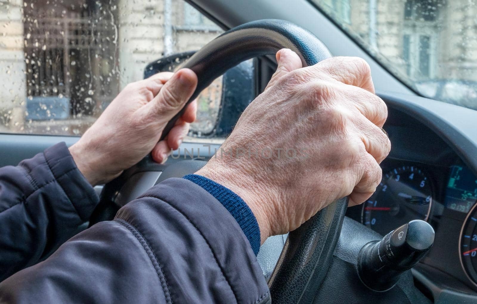 Close-up of the hand of an adult man in a passenger car, holding the steering wheel in cold rainy weather in the city. The man is driving the car.