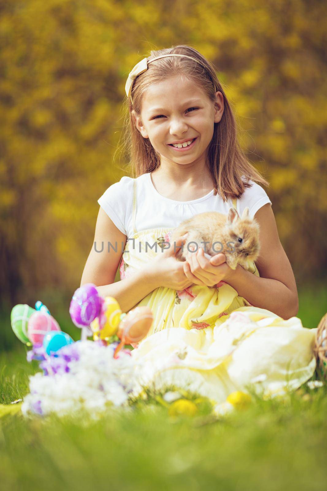 Beautiful smiling little girl holding cute bunny and sitting on the grass with Easter eggs and flowers in spring holidays. Looking at camera. 