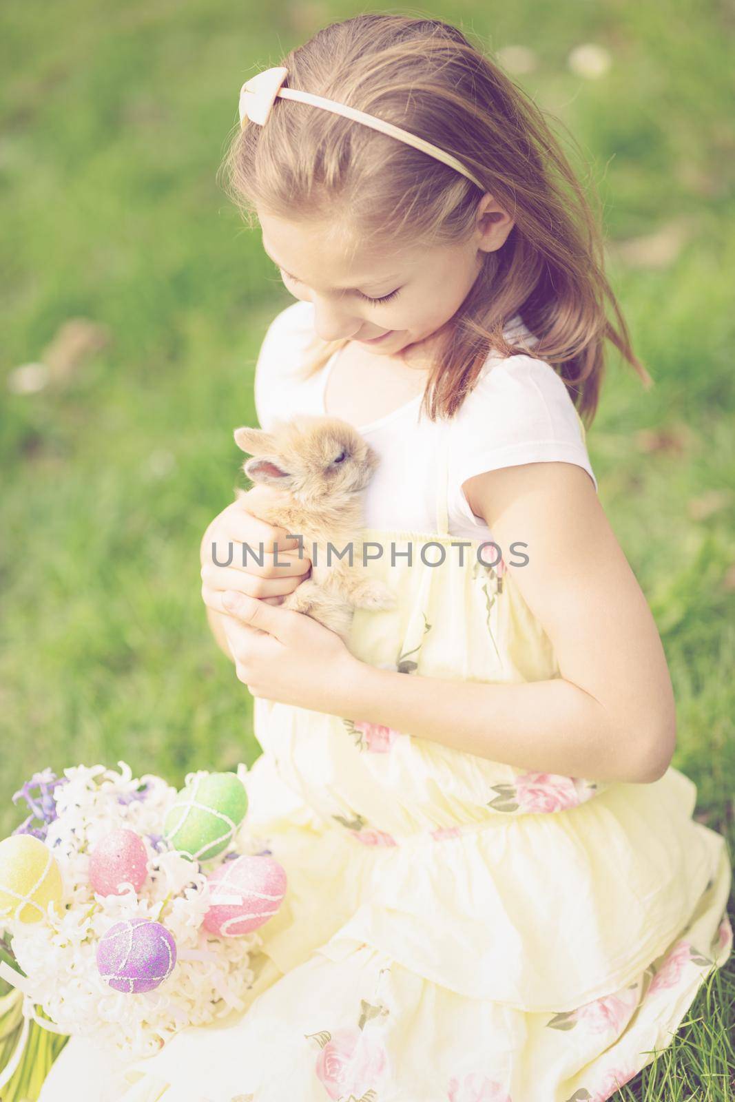 Beautiful smiling little girl playing with cute bunny and sitting on the grass with Easter eggs and flowers in spring holidays. 