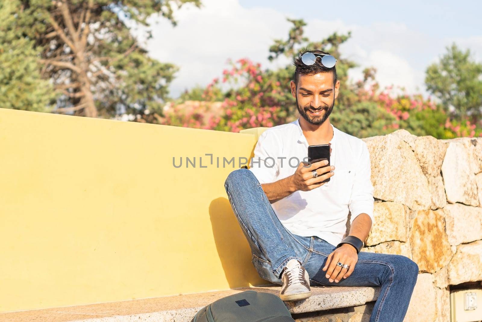 Young beautiful happy stylish guy with backpack sitting on a city bench waiting for a public transportation way to his travel destination using smartphone to sharing pictures and video of life moments