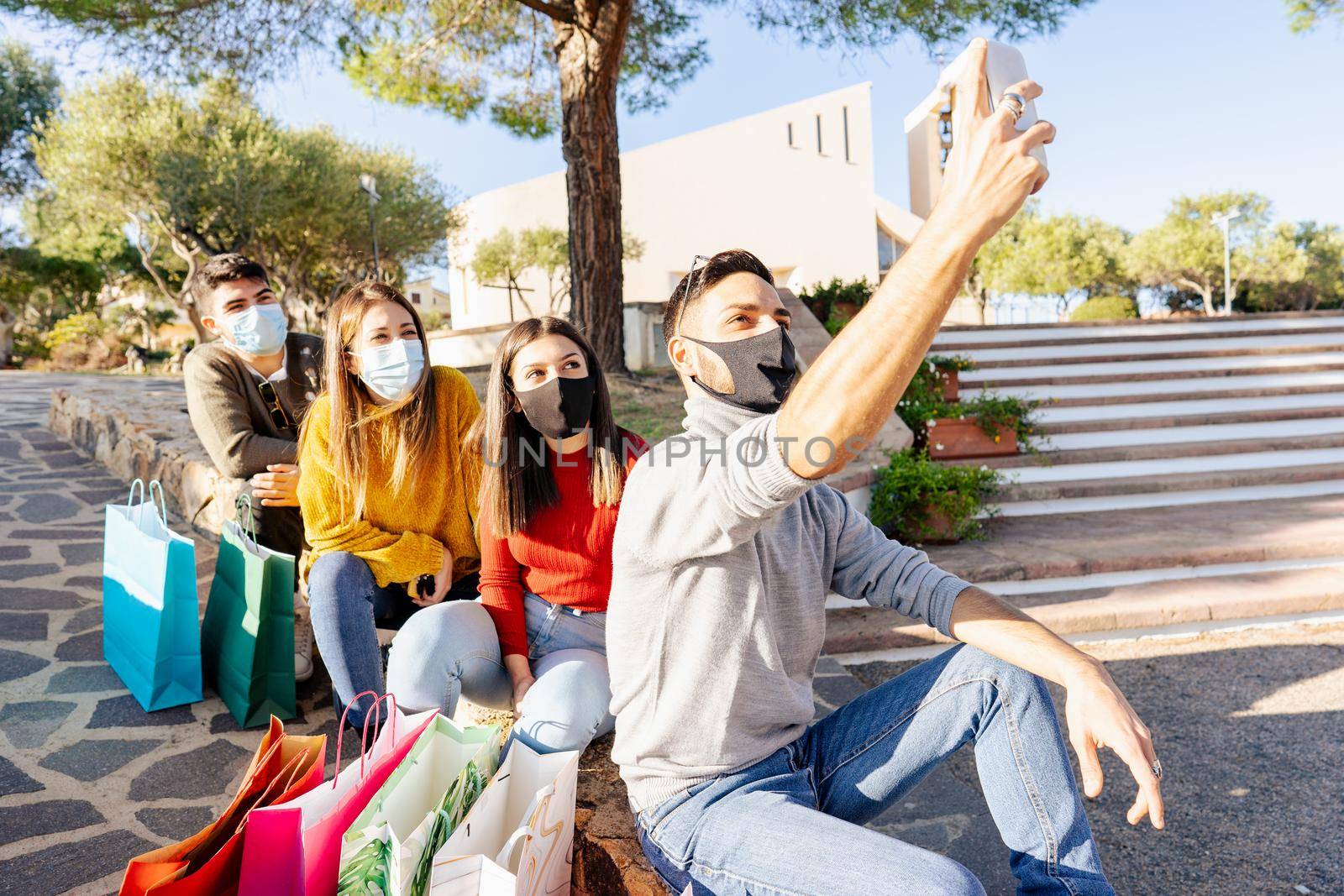 Group of young people in protective medical mask among several colored shopping bags waiting out the mall market enjoying internet mobile connection to take a selfie and sharing it on social network