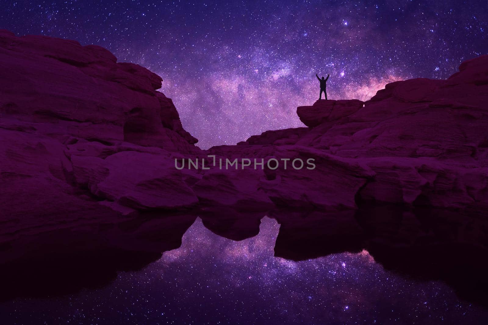 Night landscape with Milky Way. Silhouette of a standing young man with raised up arms on the mountain. Beautiful Universe. Travel background with blue night starry sky