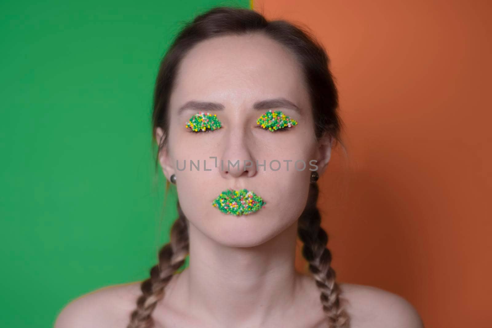 Young woman portrait with a candy makeup - multicoloured pastry topping pearls on her lips and eyelids. On a green and orange background. Easter theme.