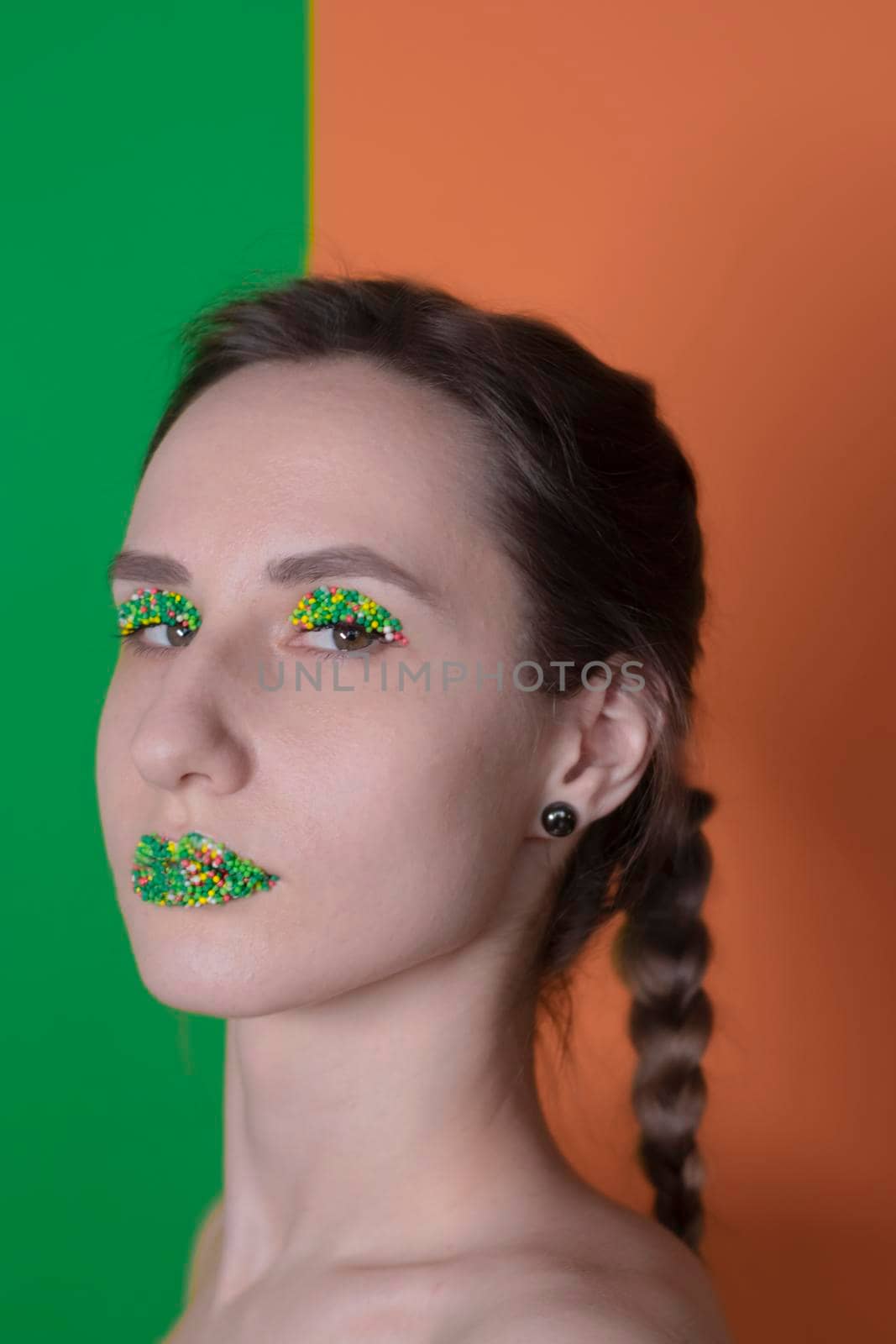 Young woman portrait with a candy makeup - multicoloured pastry topping pearls on her lips and eyelids. On a green and orange background. Easter theme.