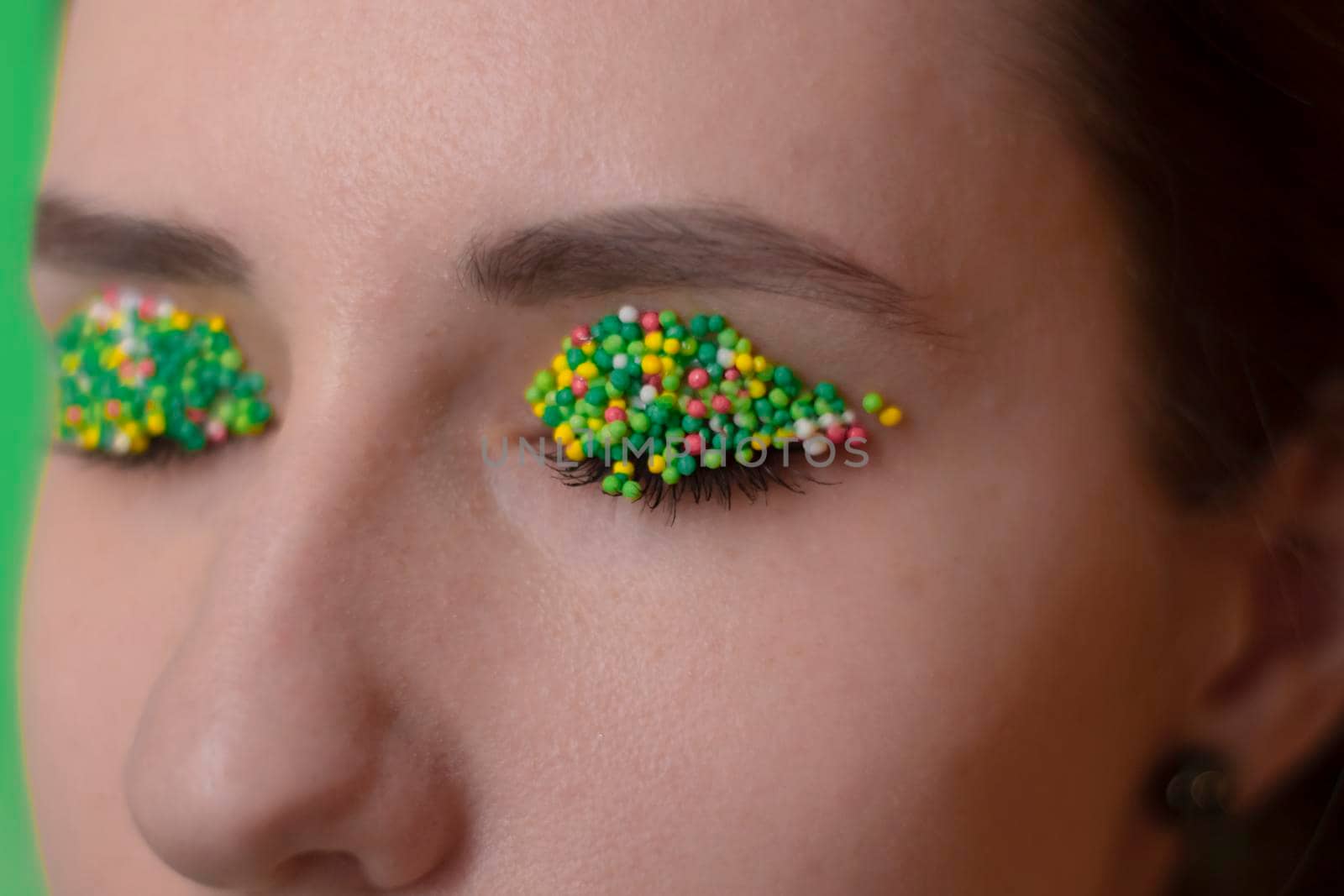 Young woman portrait with a candy makeup - multicoloured pastry topping pearls on her lips and eyelids. On a green and orange background. Easter theme. by oliavesna