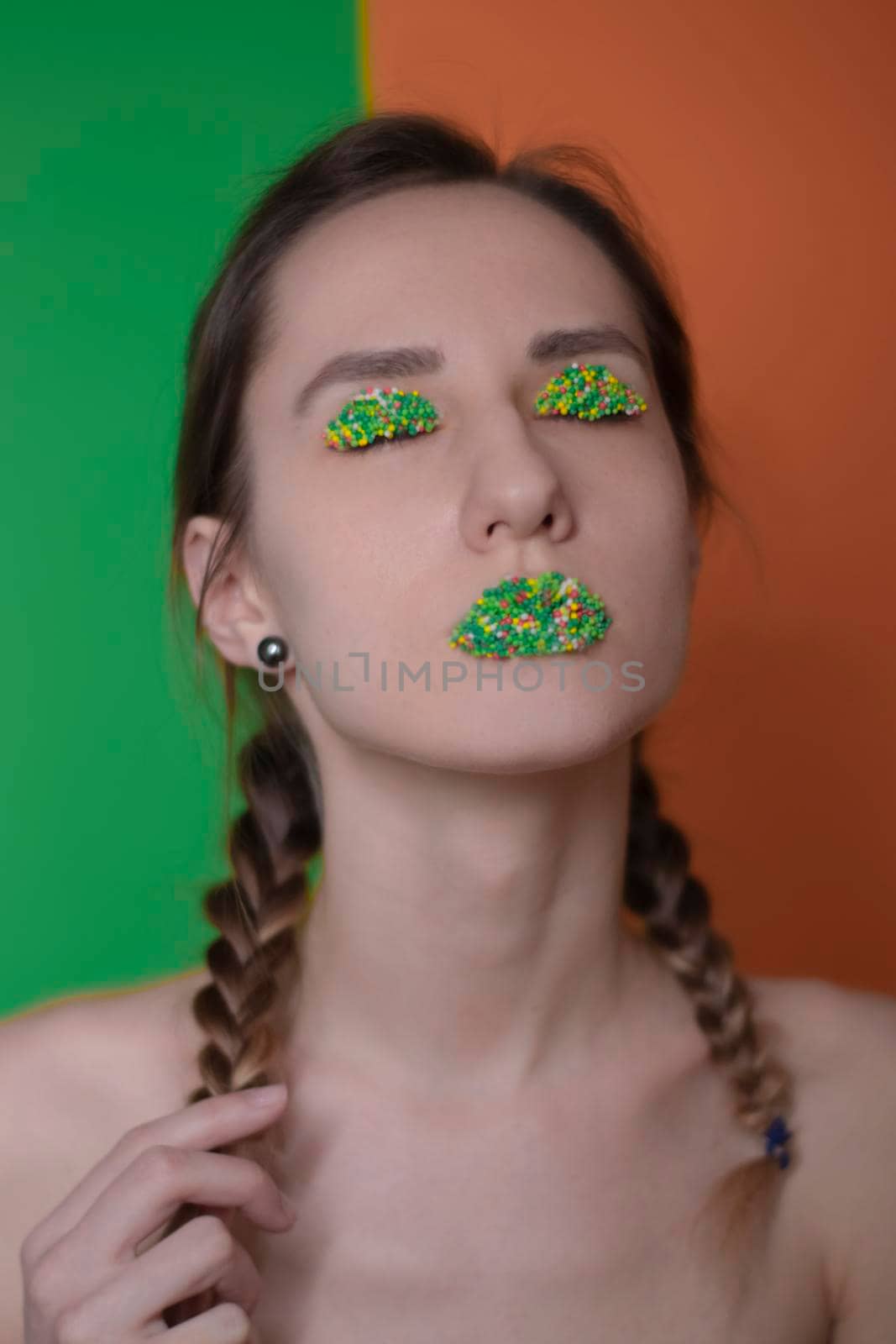 Young woman portrait with a candy makeup - multicoloured pastry topping pearls on her lips and eyelids. On a green and orange background. Easter theme.