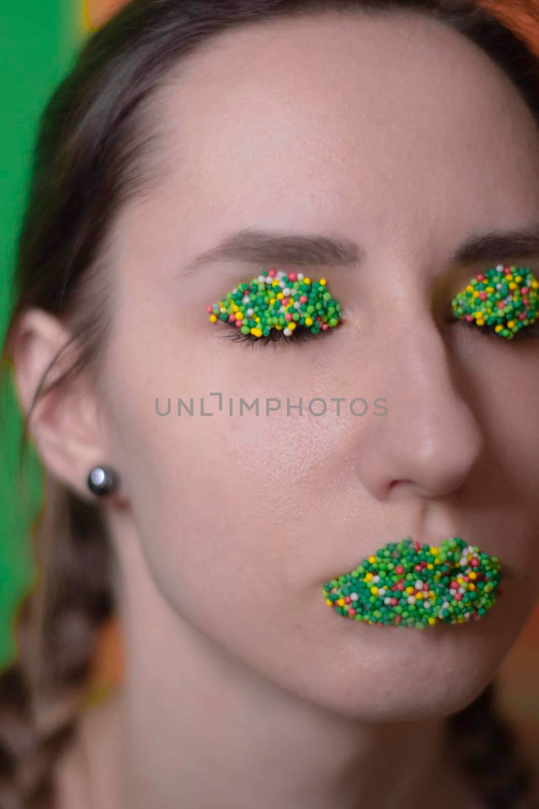 Young woman portrait with a candy makeup - multicoloured pastry topping pearls on her lips and eyelids. On a green and orange background. Easter theme.