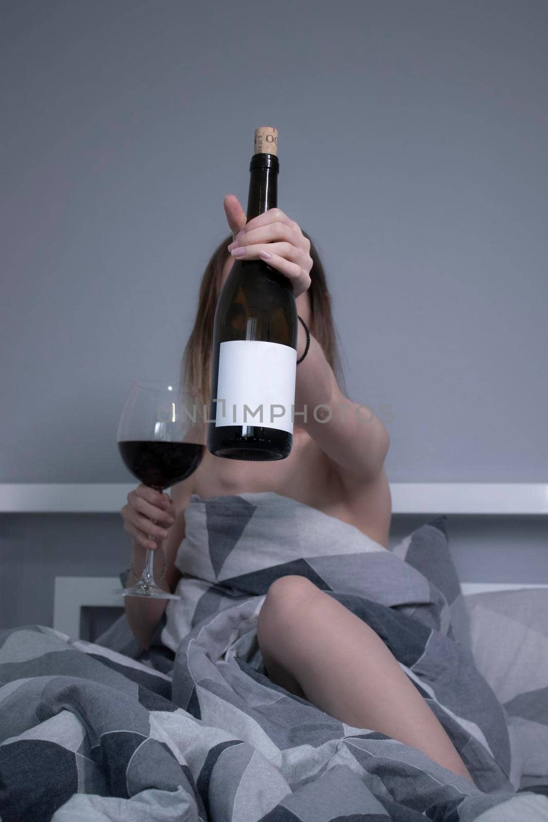 happy beautiful slender half-naked girl shows us a bottle of wine without a label, in bed with gray sheets in triangles by oliavesna