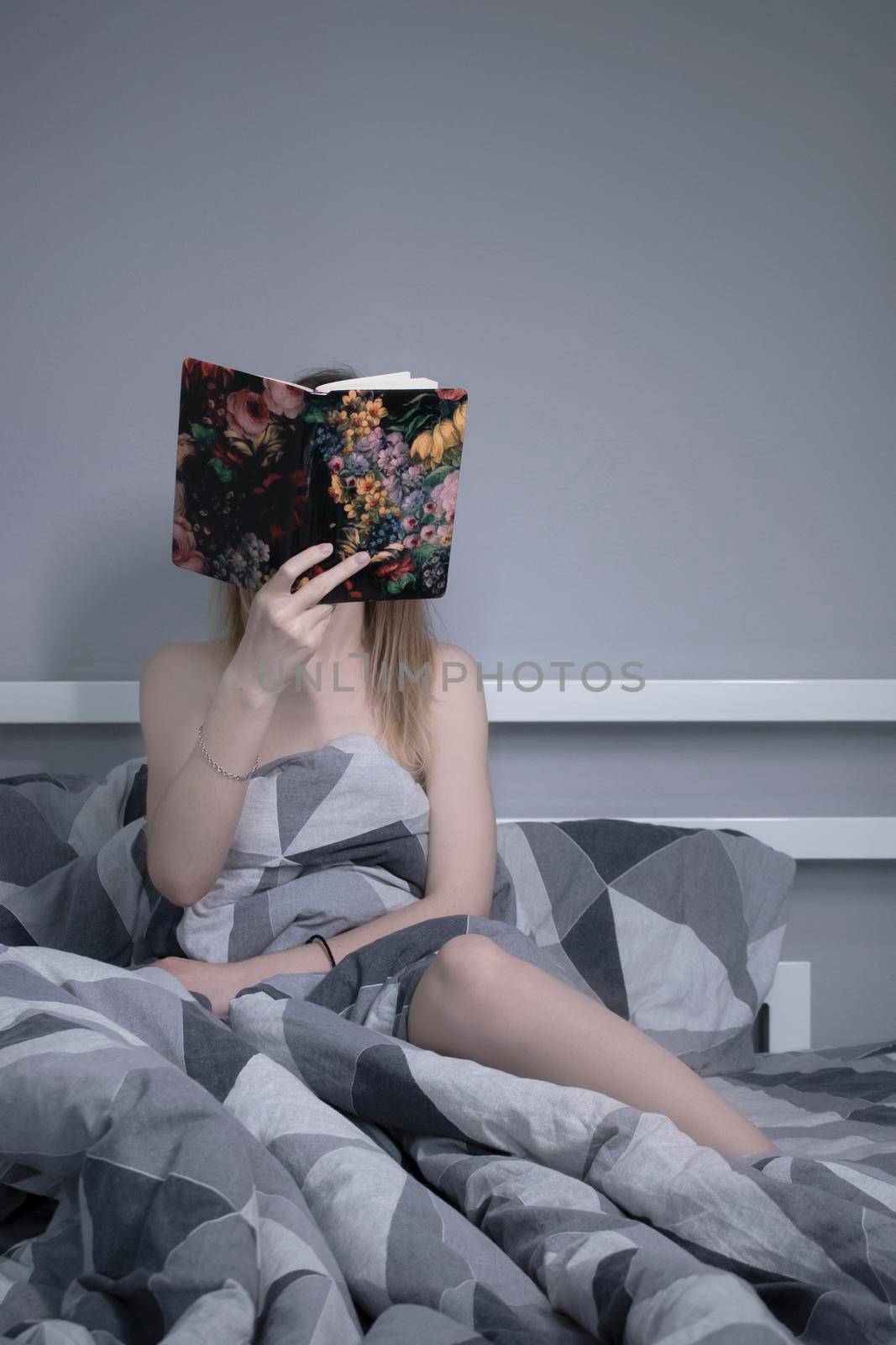 Half-naked slender blonde girl reads a beautiful book in flowers in bed with grey sheets in triangles by oliavesna