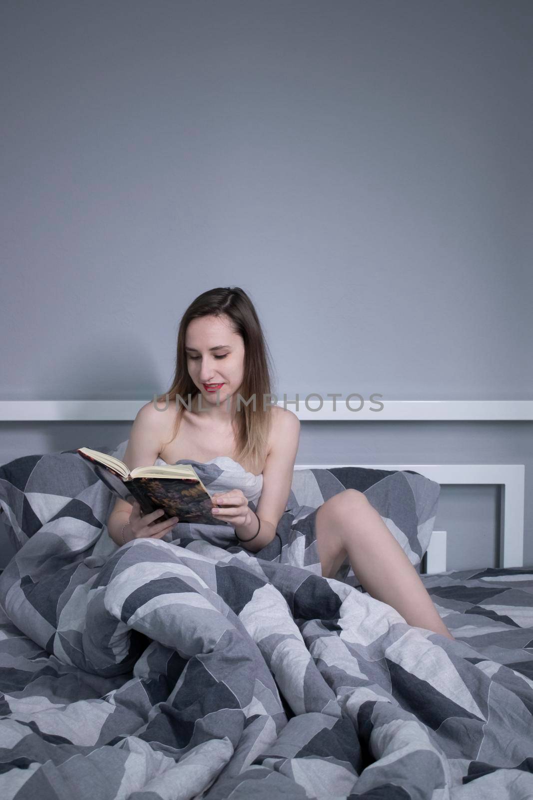 Half-naked slender blonde girl reads a beautiful book in flowers in bed with grey sheets in triangles - vertical photo