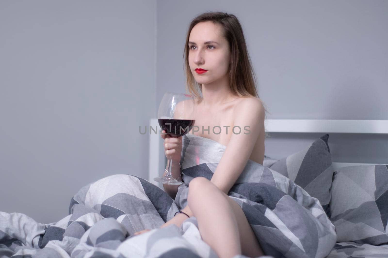 happy beautiful slender half-naked girl holds a huge glass of red wine in bed with gray sheets in triangles - vertical photo