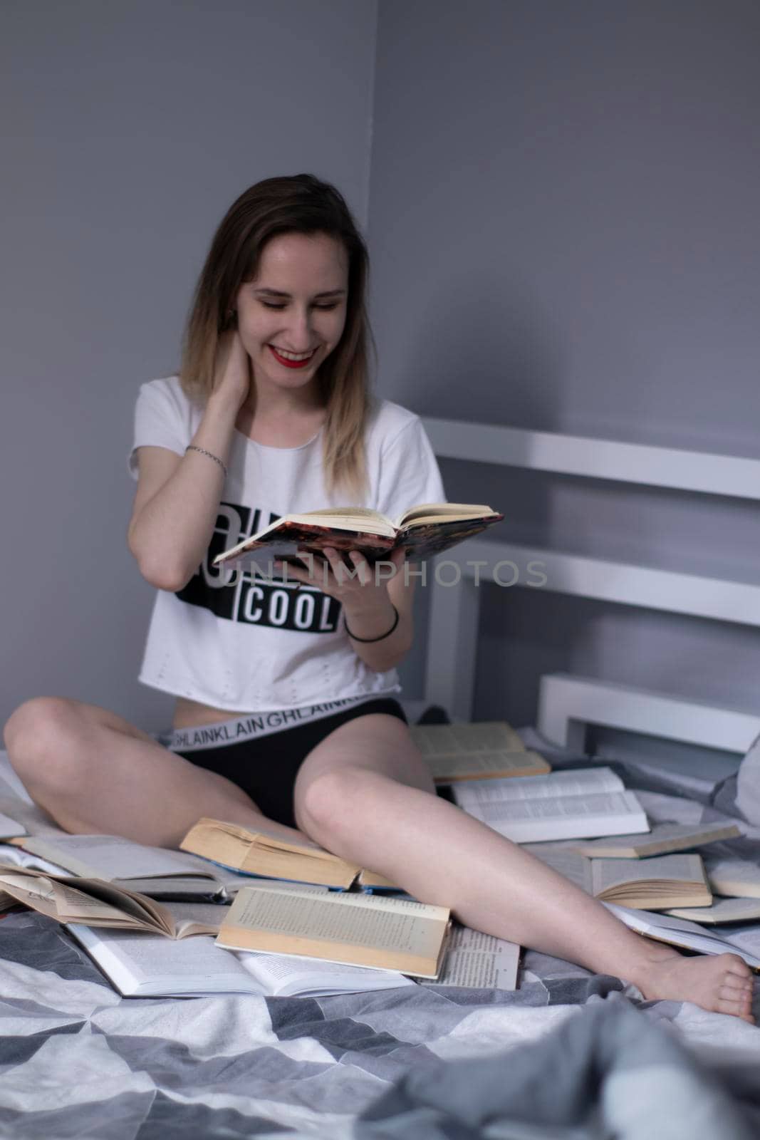 happy beautiful slim girl sitting on the bed with a pile of books, reads. Home schooling. Studying in quarantine. Reading is helpful. Vertical photo