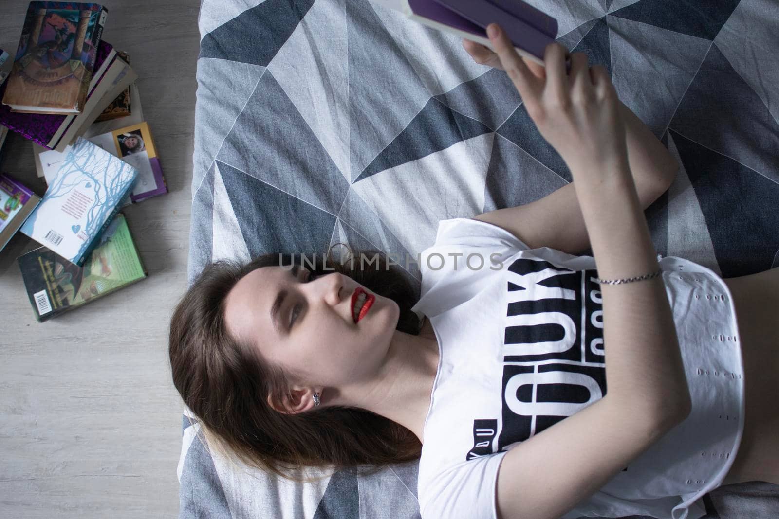 happy beautiful slim girl reading a book lying on the bed with a pile of books nearby. Grey bedroom and sheets. Homeschooling. Studying in quarantine. Reading is helpful. horizontal photp