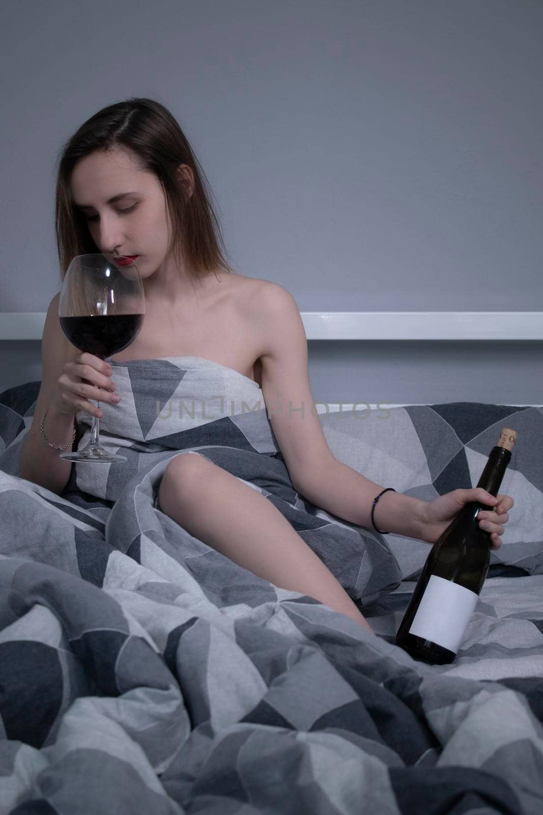 beautiful slender half-naked girl drinks red wine from a huge glass and a bottle with no label a in a bed with gray sheets - vertical photo