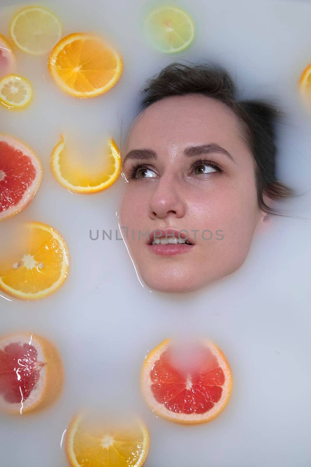 Woman portrait in milk bath with oranges, lemons and grapefruits. Healthy dewy skin. Fashion model girl, spa and skin care concept. Spring colours - yellow, orange, red.