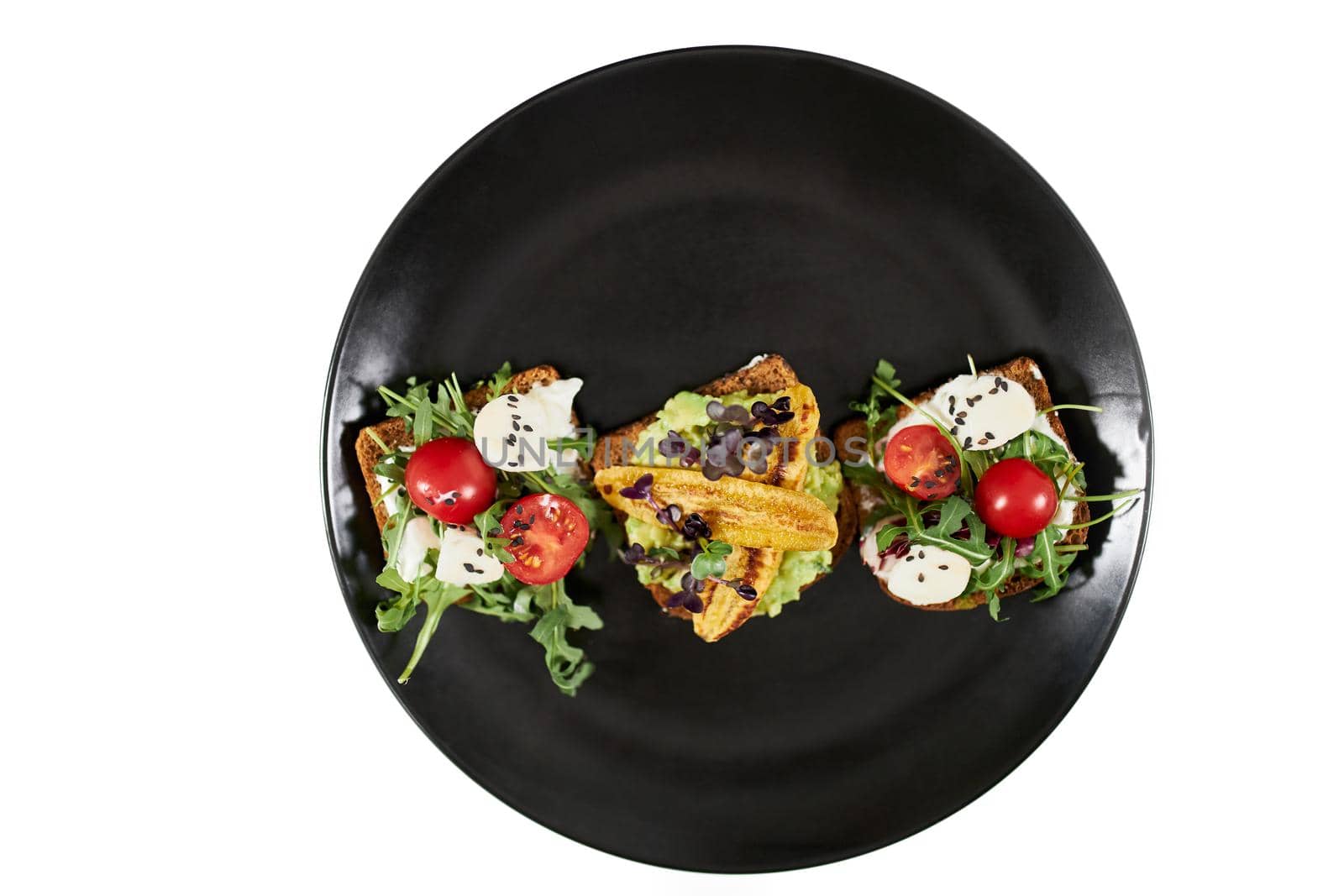 Top view of beautiful modern black plate with appetizing toasts with tomatoes,mozzarella and arugula on white background. Concept of healthy and tasty snacks at home.