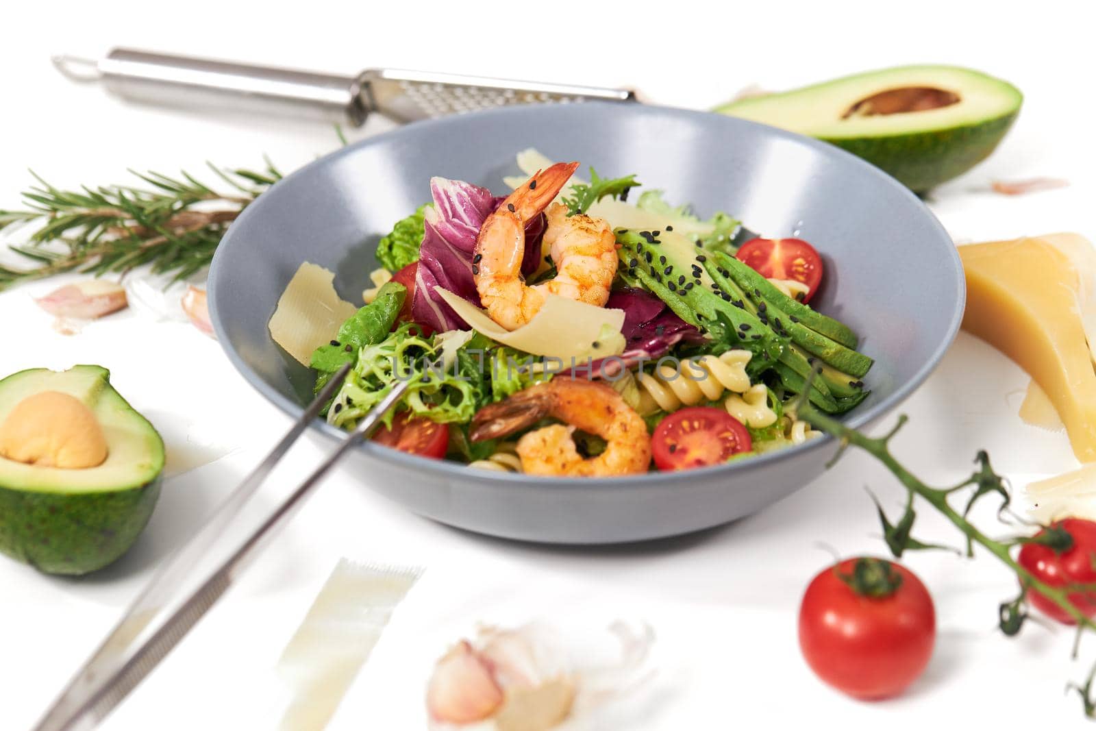 Close up of modern blue plate with vegetables salad with pasta,avocado and shrimps on white background. Concept of process cooking healthy and appetizing salad at home.