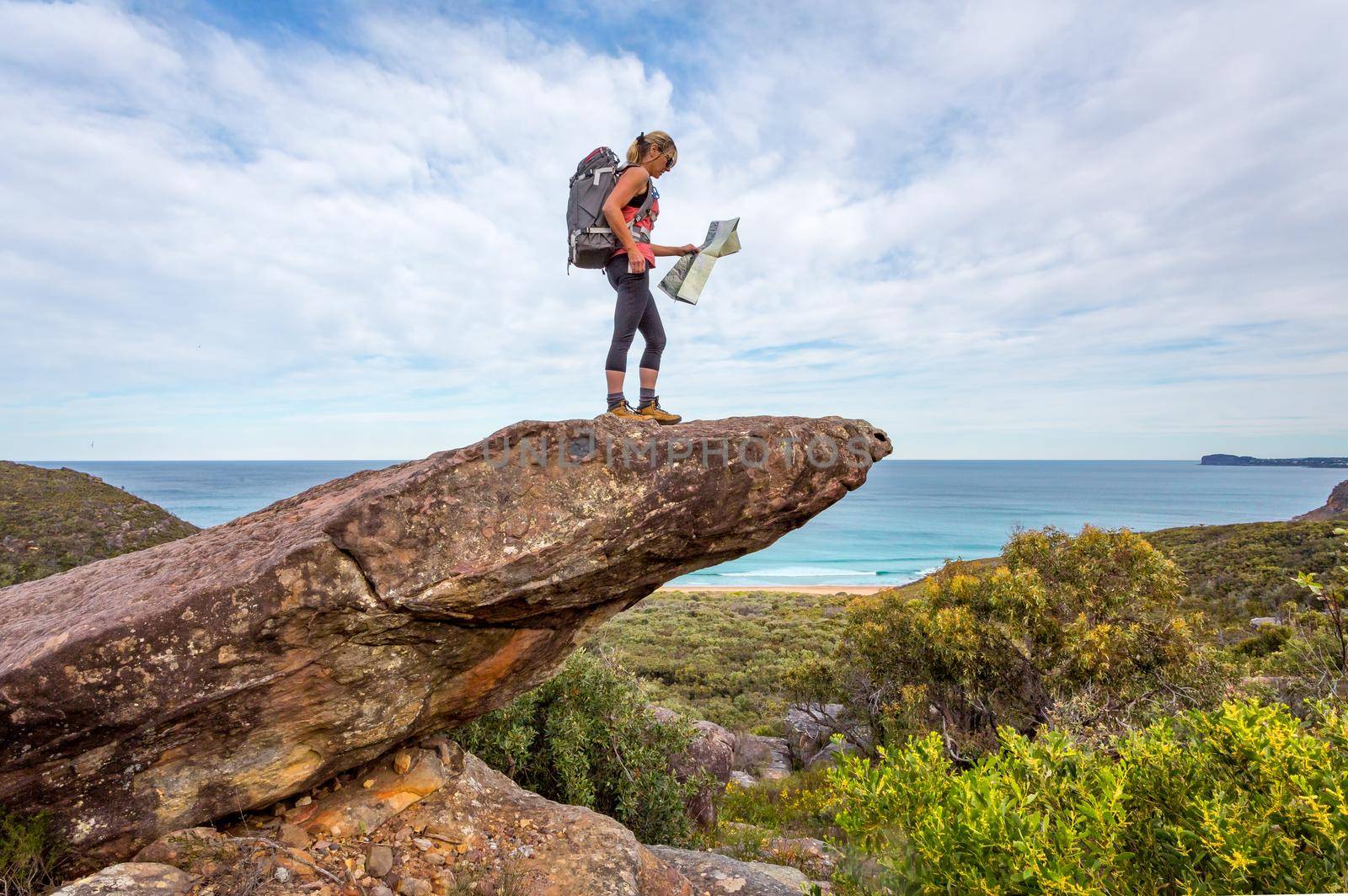 Female hiker stands on a rock with coastal views, she is carrying a backpack and holding a map