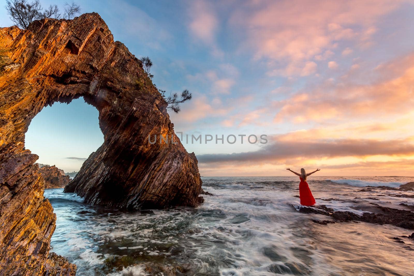 Woman in red dress standing in ocean waves by a sea arch by lovleah