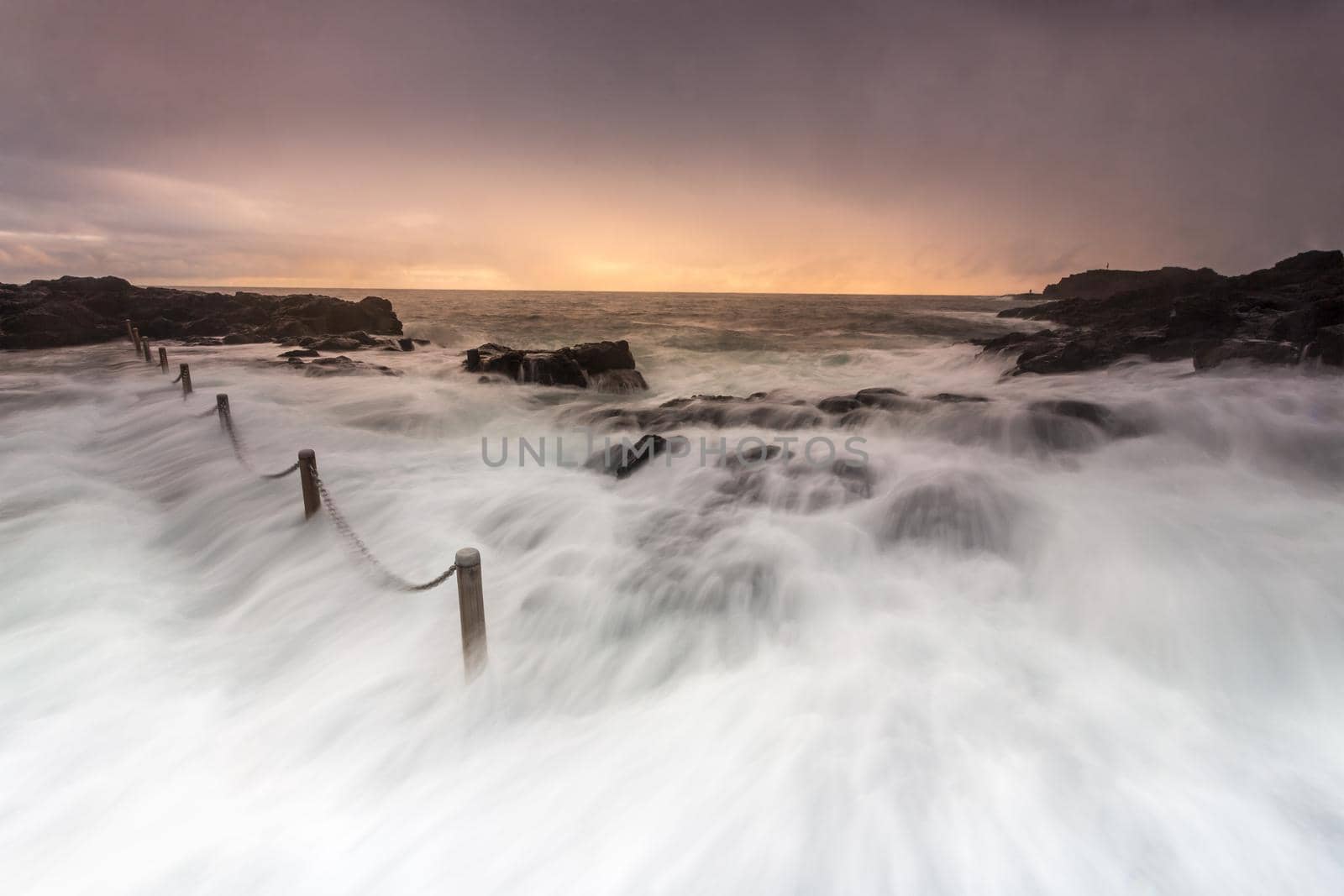 Large waves wash over rocks and into rock pool.  The heavy chains are in motion and so is the water and a few loose rocks as they toss with the water.  Sun breaks through some thinner cloud in the early morning adding some soft warmth.