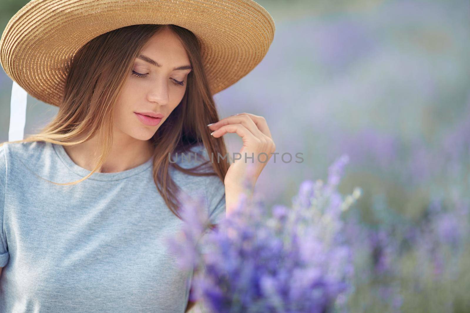 Stunning young woman sitting at table with lavender violet bouquet. Pretty fashionable girl wearing straw hat and blue dress posing in lavender field. Concept of nature, beauty, fashion.