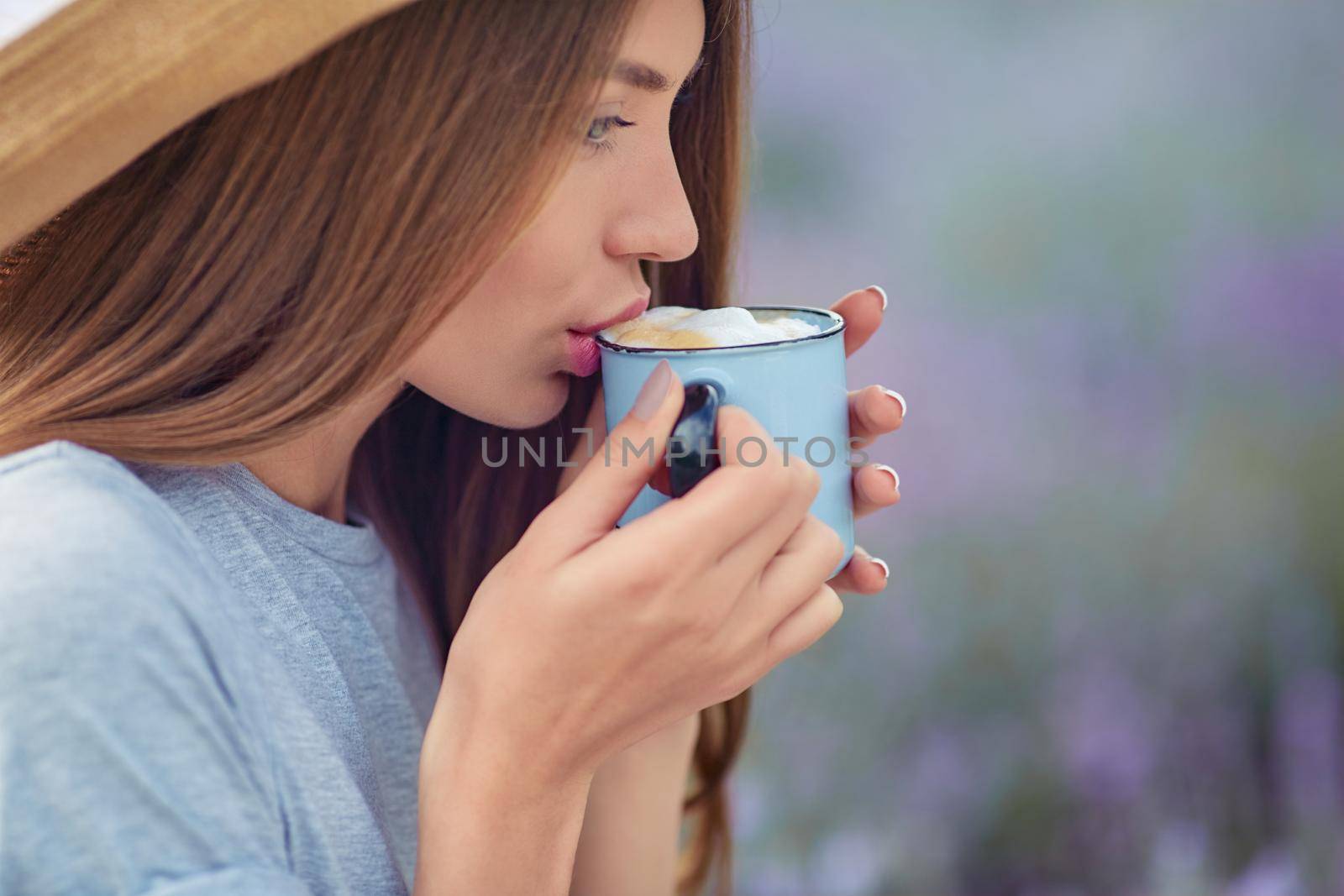 Side close up view of young girl wearing straw hat enjoying hot drink outdoors. Stunning woman drinking coffee with foam, posing in lavender field. Concept of beauty, nature, drink.