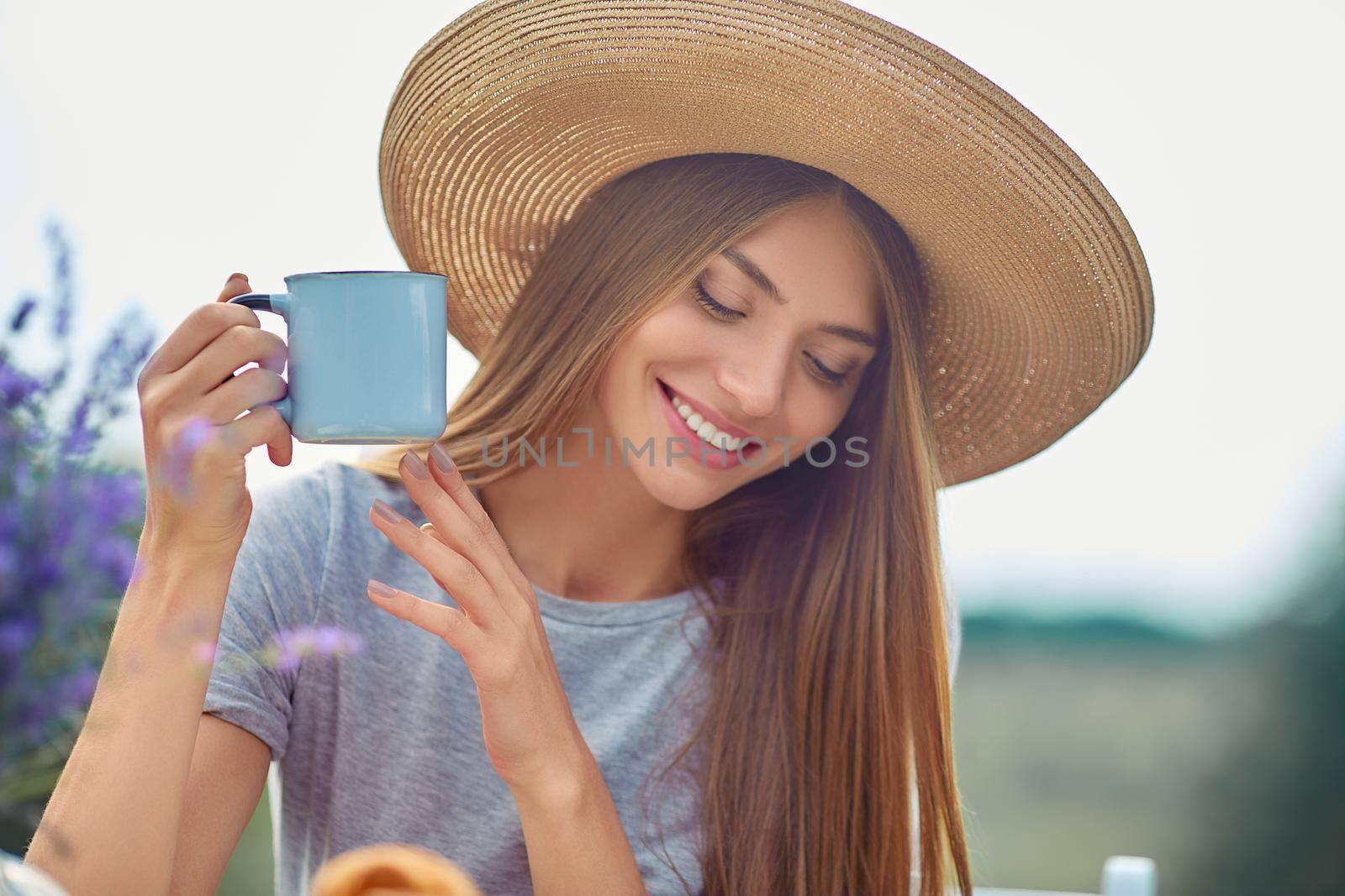 Selective focus of young girl wearing big straw hat and dress holding hot drink outdoors. Stunning caucasian woman drinking coffee, posing in lavender field. Concept of beauty, nature, drink.