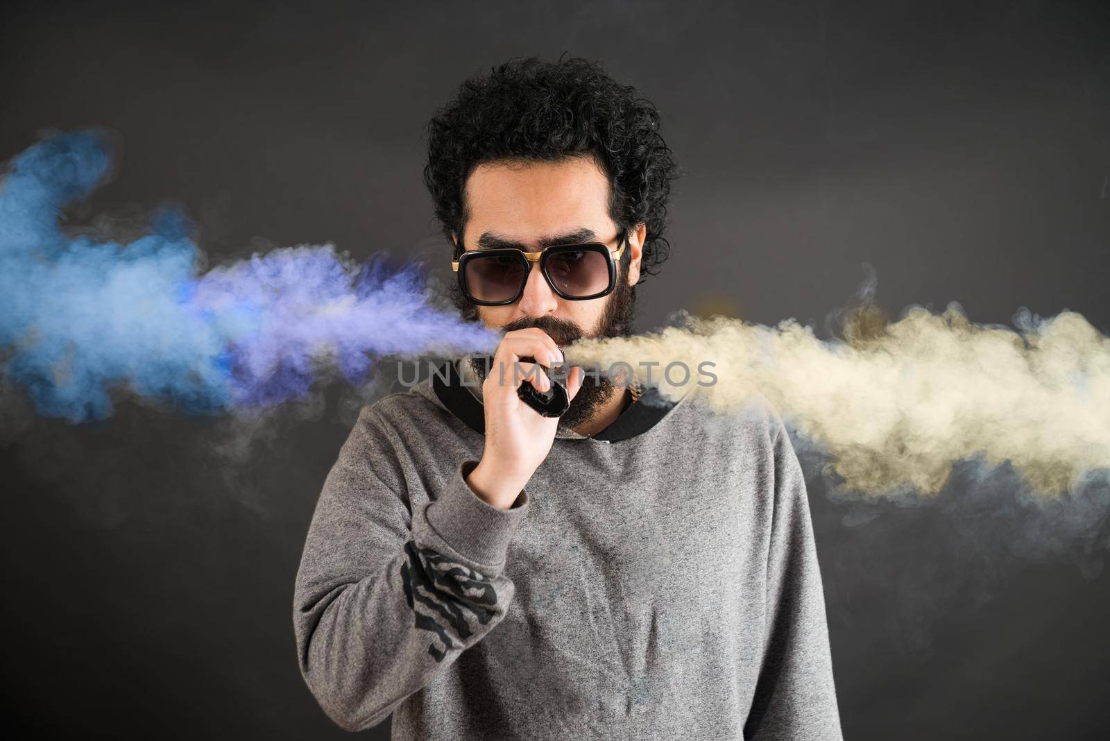 A young man smokes a vape and exhales a blue and yellow smoke on a black background.