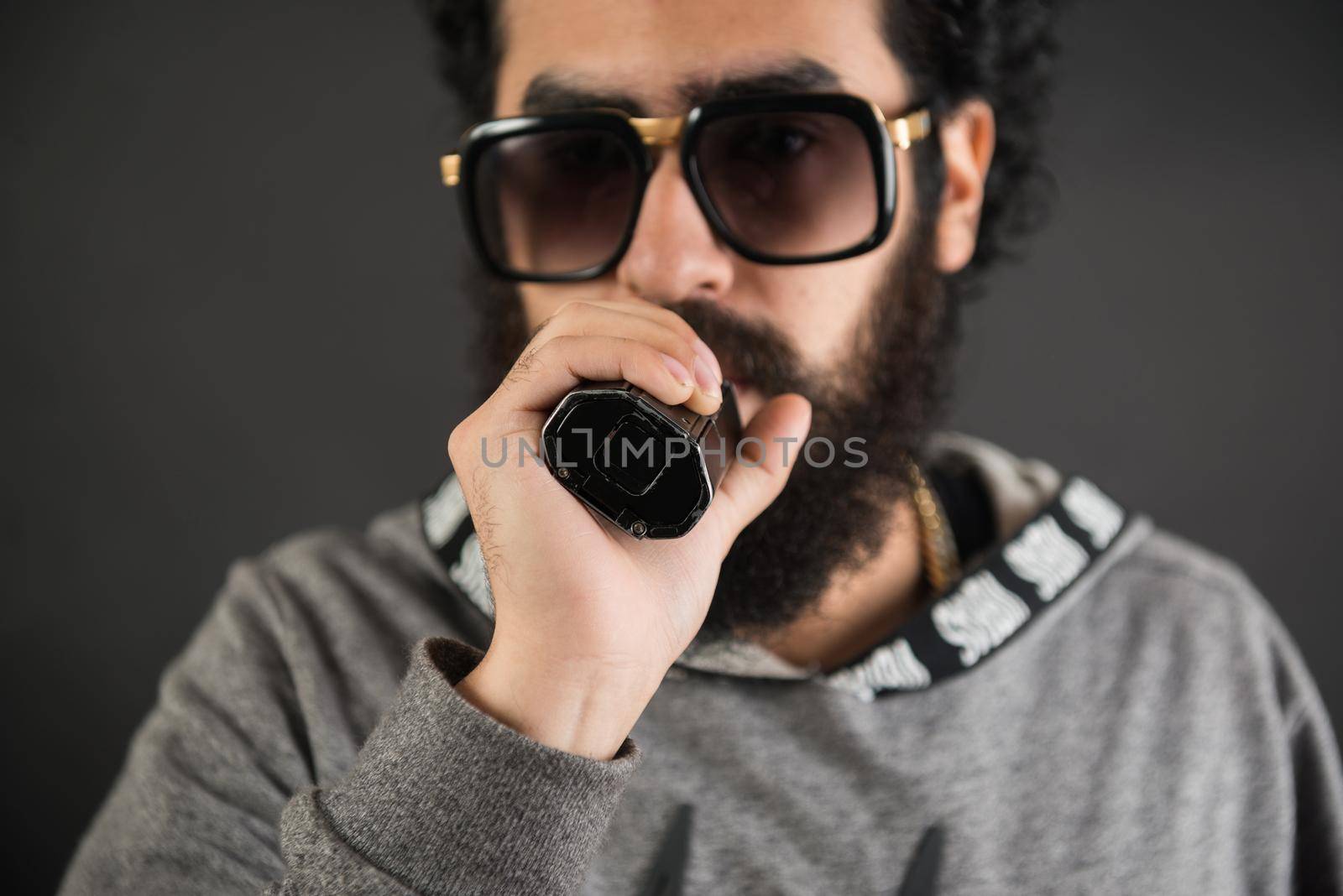 Portrait of a young guy with big beard and sunglasses and vaping an electronic cigarette on a black background.