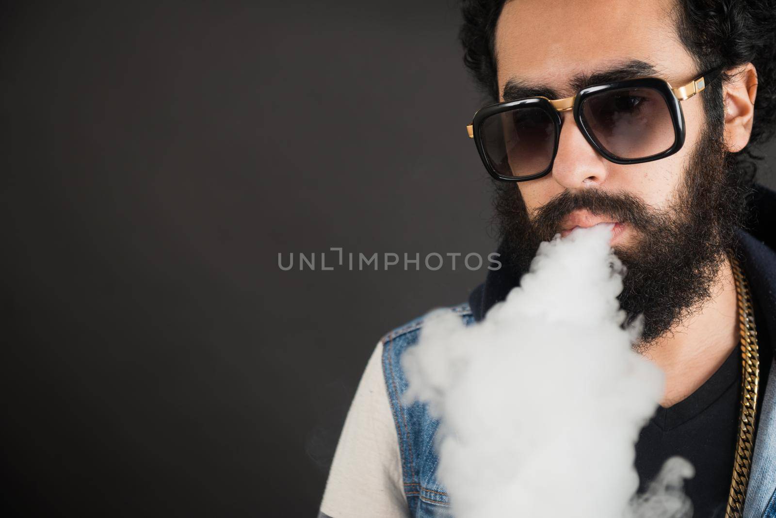 Young man vaping, studio shot. Bearded guy with sunglasses blowing a cloud of smoke on black background. Concept of smoking and steam without nicotine, copy space