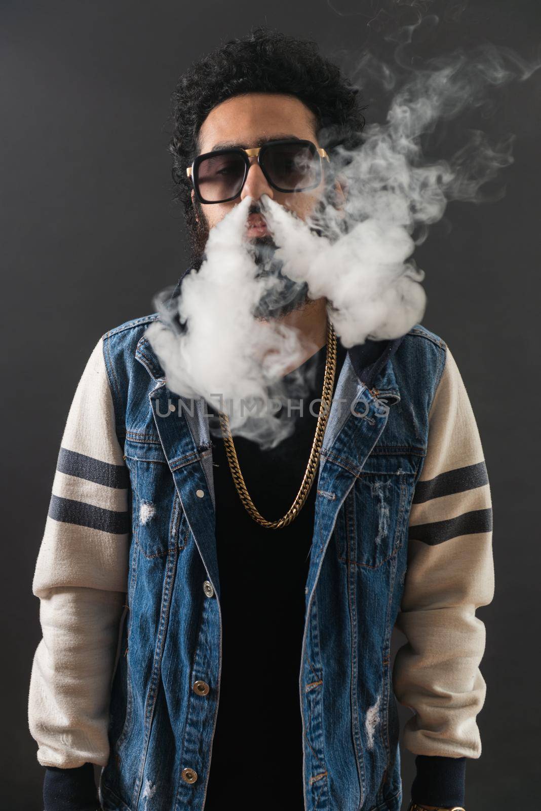 Young man vaping, studio shot. Bearded guy with sunglasses blowing a cloud of smoke on black background. Concept of smoking and steam without nicotine, copy space