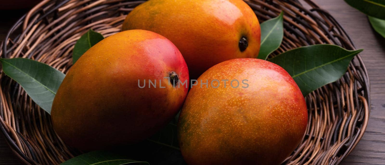 Mango. Close up of fresh ripe mango fruit on a bamboo sieve over dark wooden table background with green leaves.