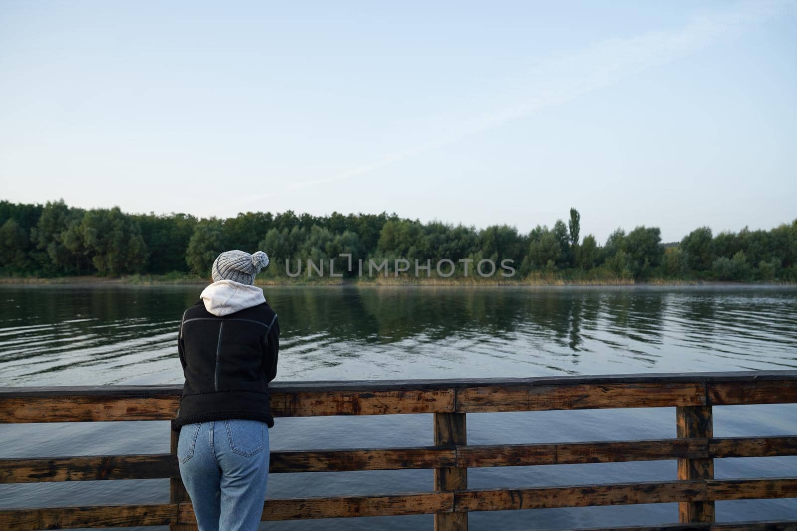 Back view of young woman in warm clothes leaning on handrails on wooden pier and joying amazing view of beautiful nature. Concept of relaxation and leisure time.