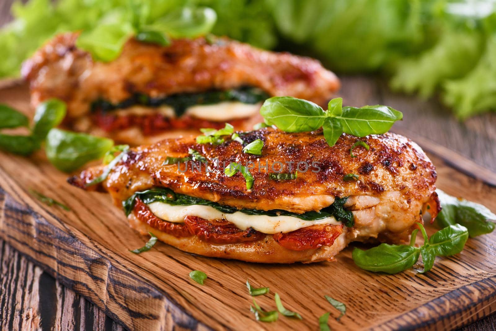 Baked chicken breast with mozzarella, sun-dried tomatoes, spinach. The meat is served on a wooden board with basil and lettuce.