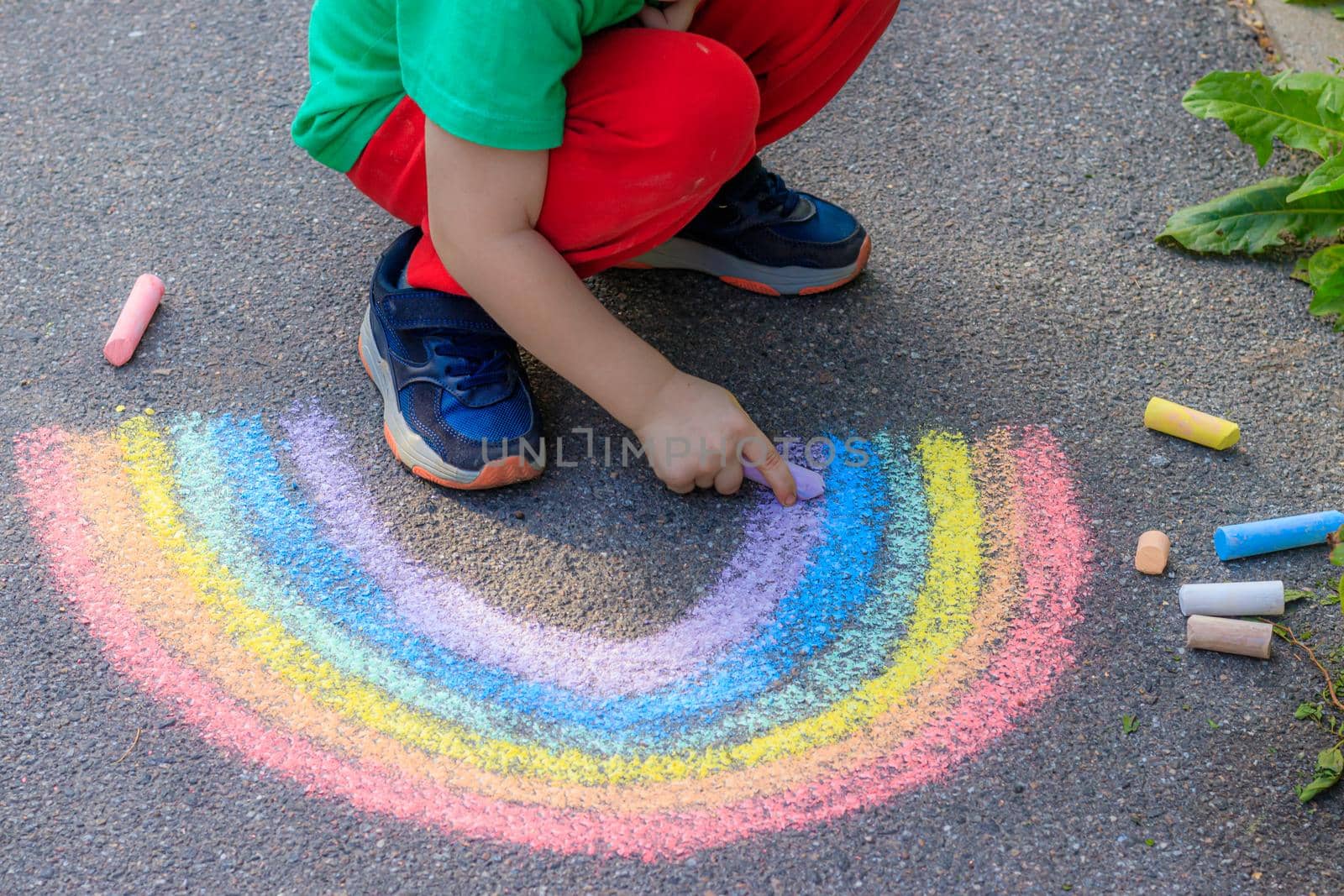 A boy draws a rainbow on the asphalt with crayons . A child draws on the asphalt . Children's drawing. Walking in the city with a child. Summer in the city. Crayons for drawing. Article about childhood.